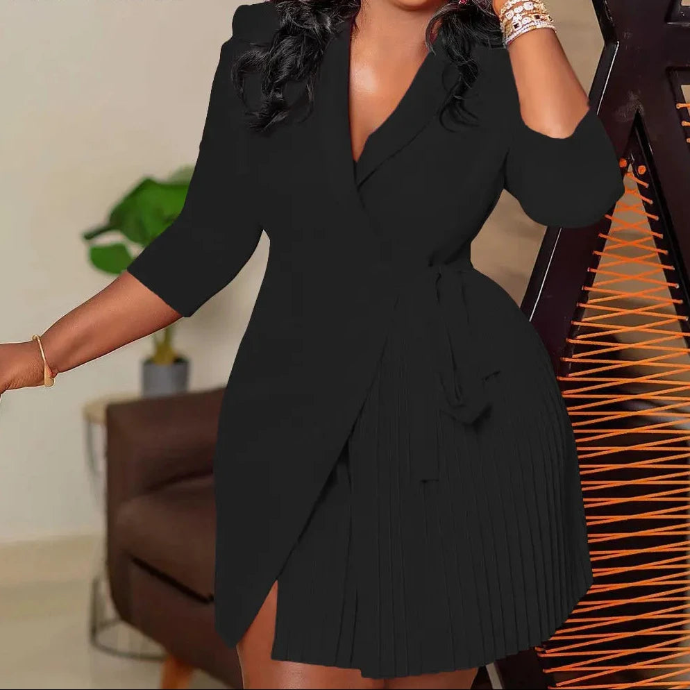 A woman wearing a black wrap-style mini dress with three-quarter sleeves and a pleated skirt, posed indoors next to a decorative chair. The brand name 'CM.