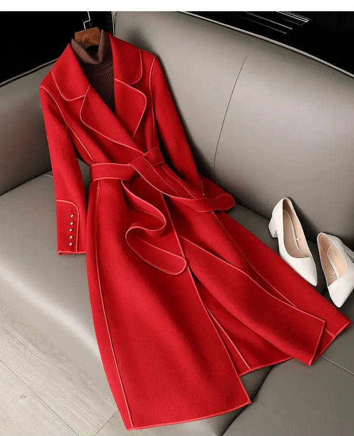 A bright red long coat with contrast light piping and a matching waist tie, displayed on a gray sofa with white pointed shoes beside it.